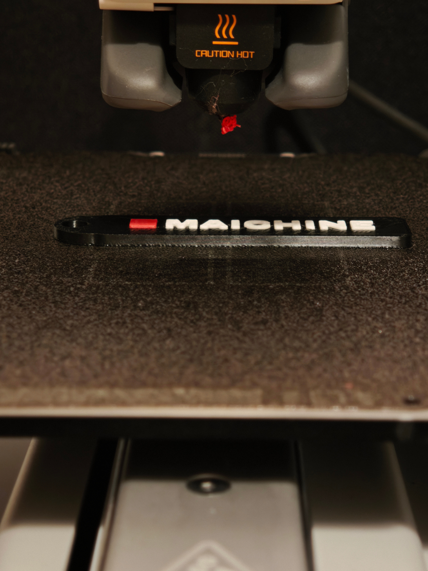 3D printer extruding a Maichine branded keychain in black, white and red filament